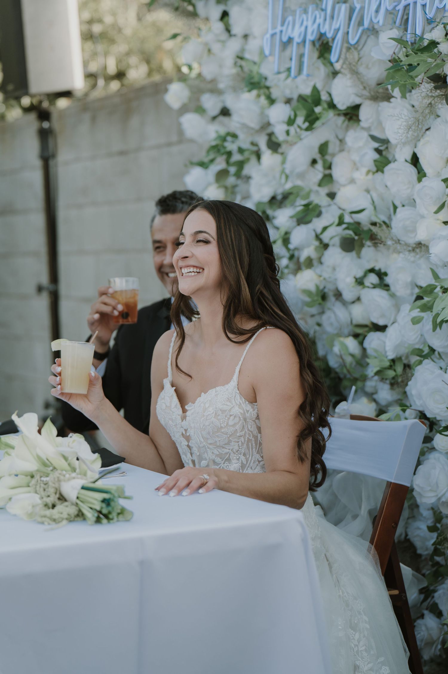 Smiling bride holding a drink at sweetheart table adorned with white floral backdrop at wedding reception.