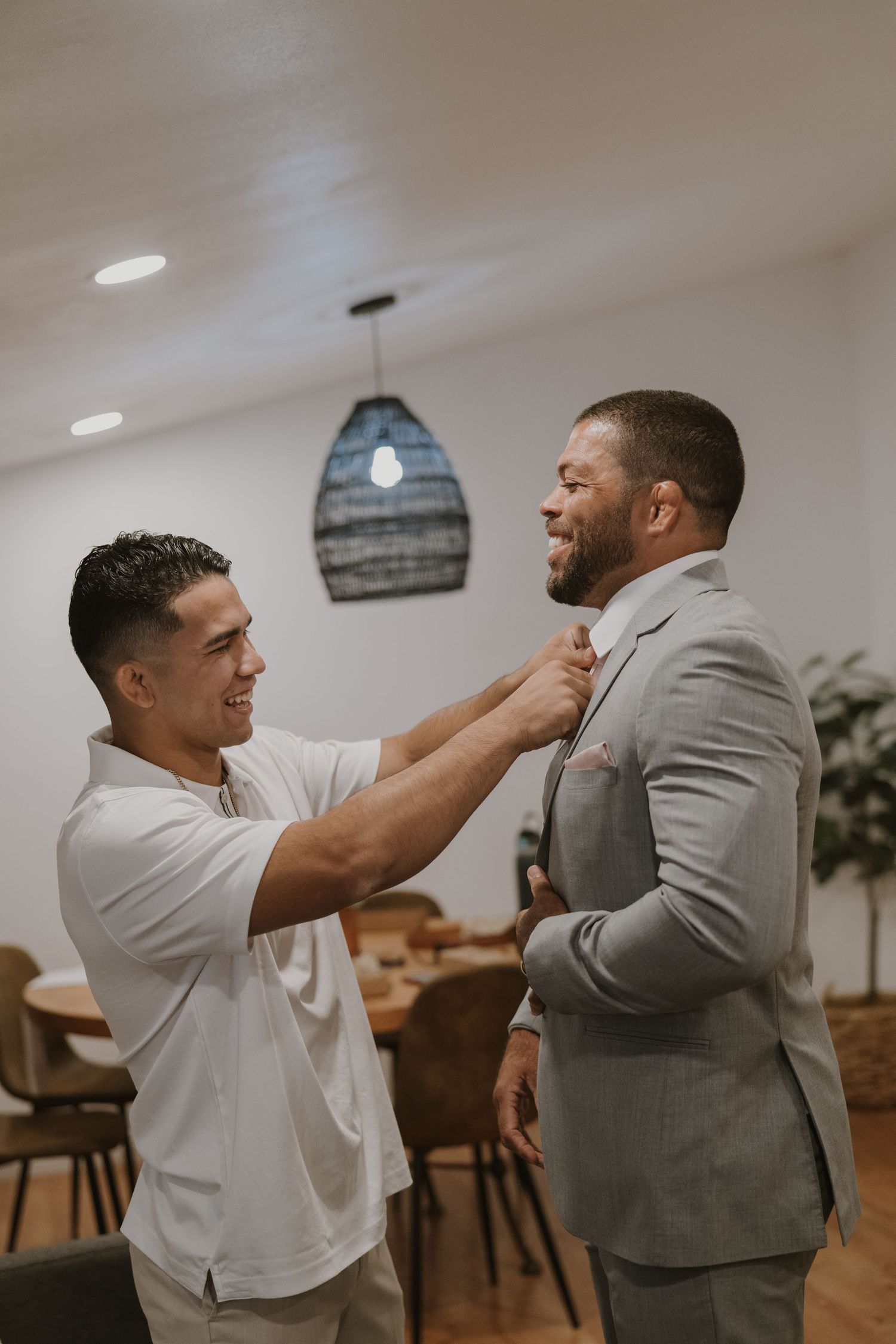 Two men smiling indoors, one in a white shirt adjusting the tie of the other in a gray suit, preparing for a formal event.