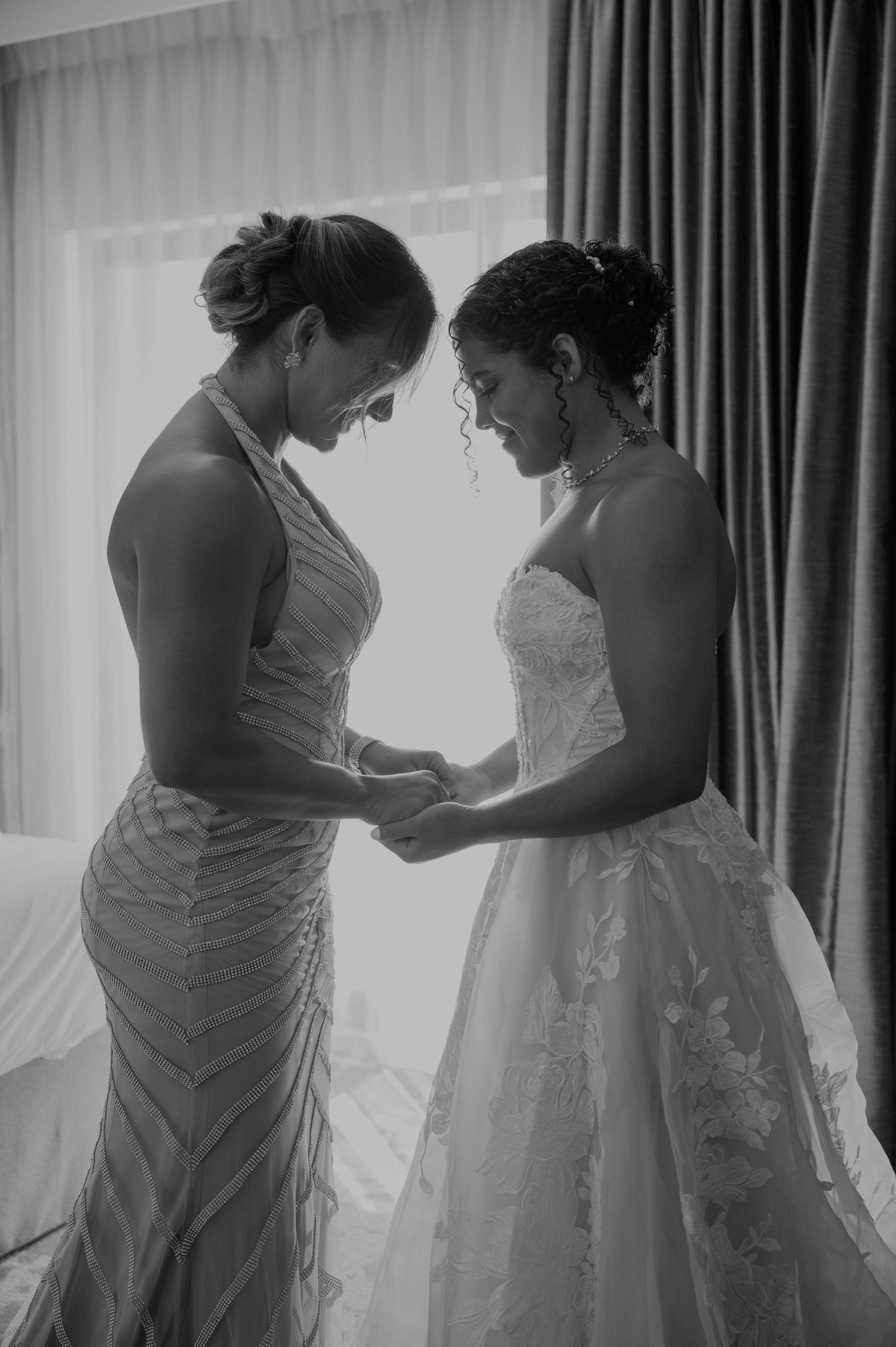 Black and white photo of bride in lace gown holding hands with bridesmaid in halter dress near window.