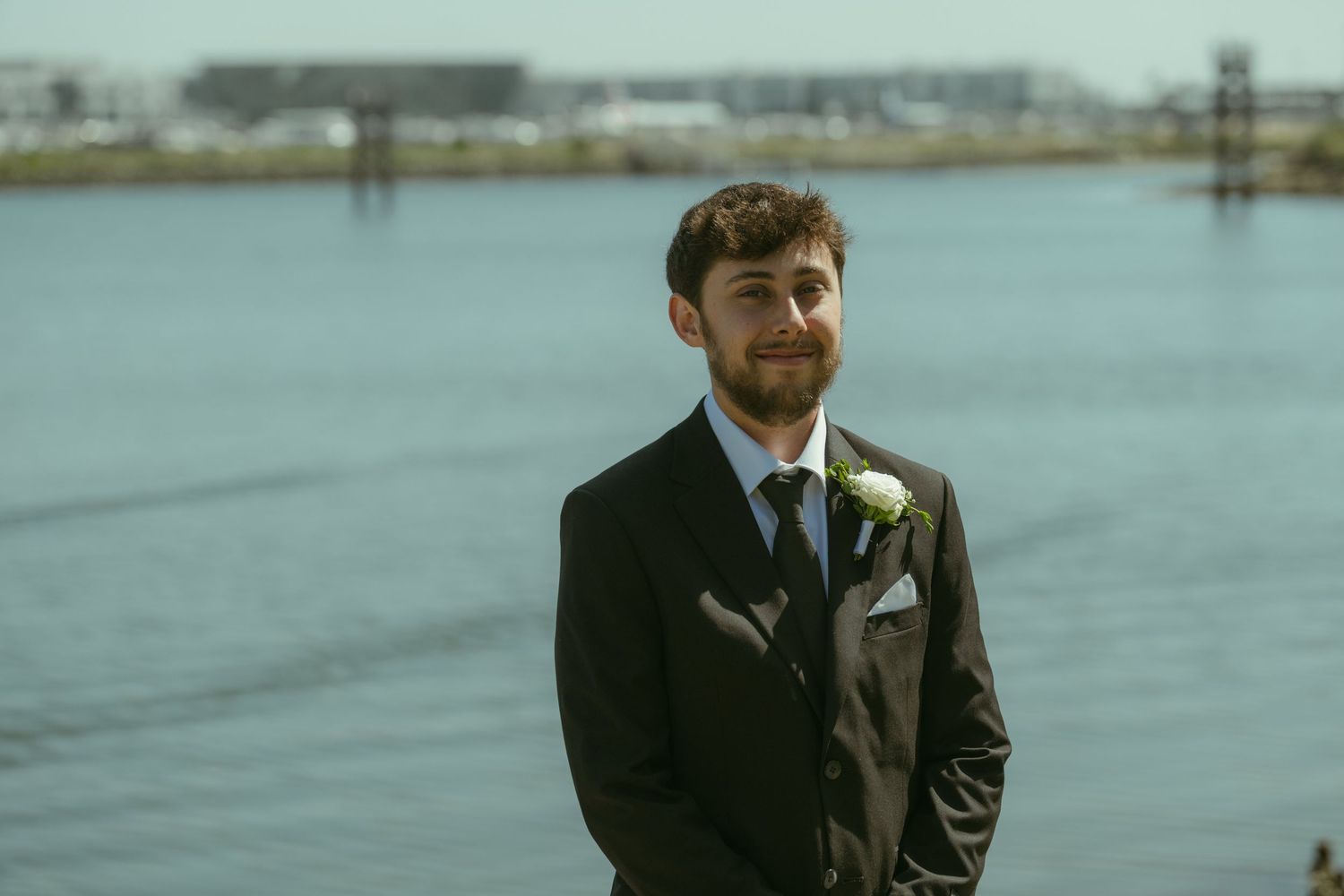 Smiling groom in a dark suit with boutonniere stands by waterfront on his wedding day.