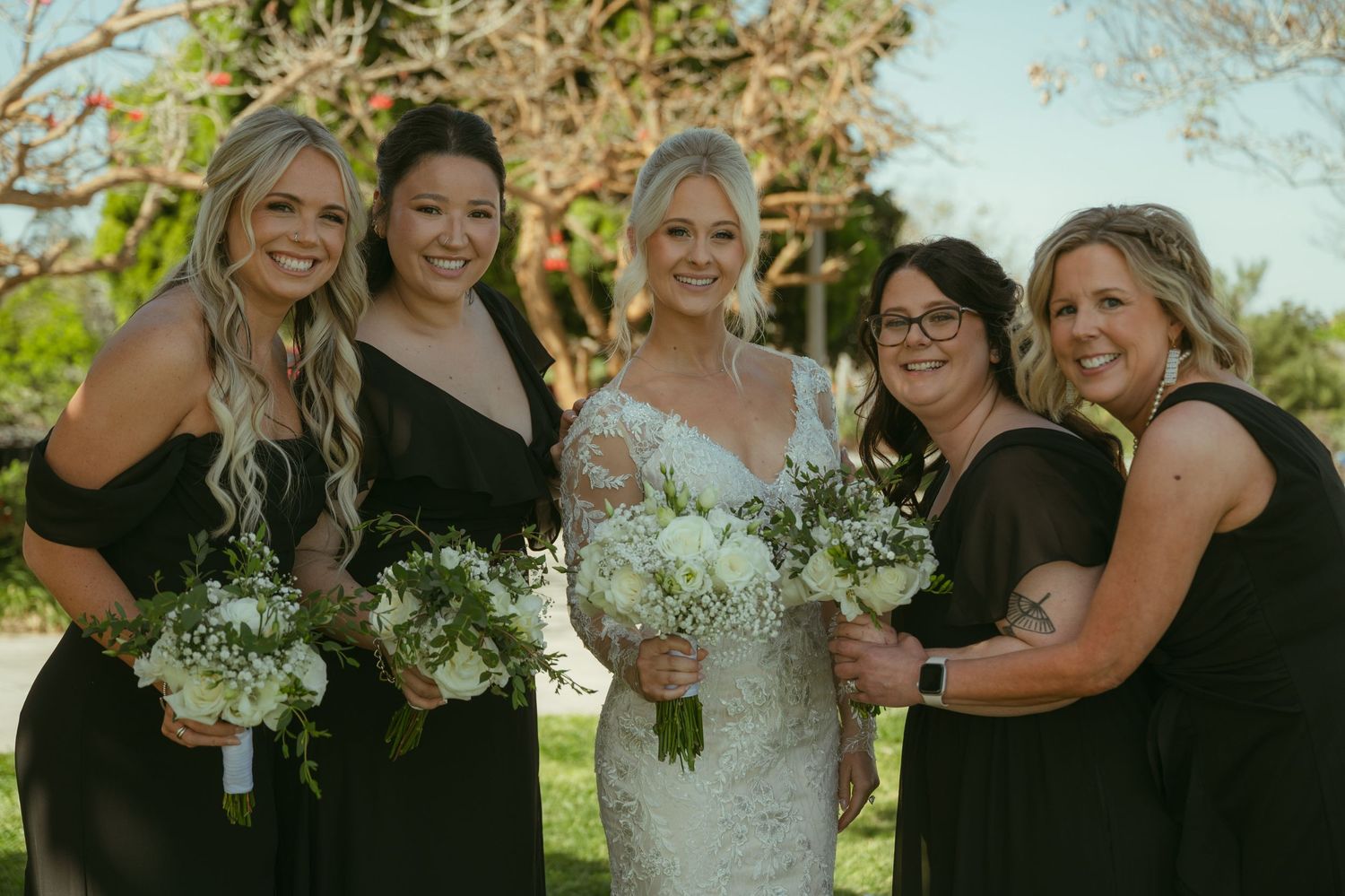 Bride in lace gown with four bridesmaids in black dresses holding white bouquets outdoors.