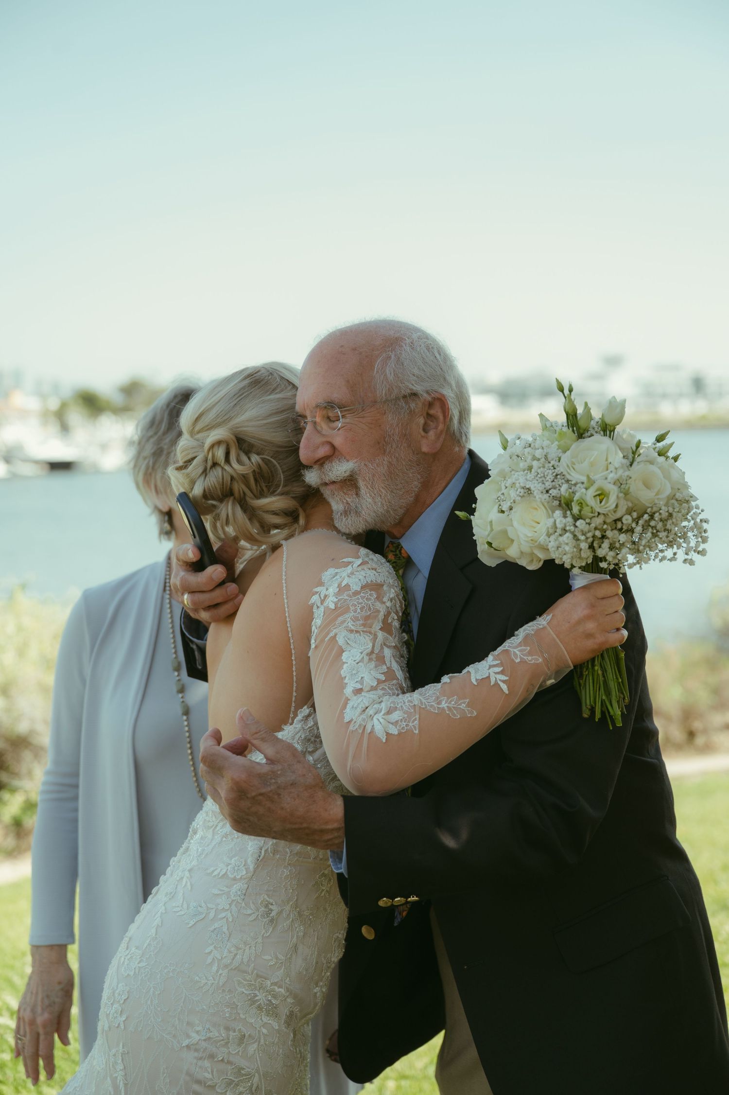 Elderly man embracing bride in lace wedding gown outdoors, bride holding white floral bouquet near waterfront.