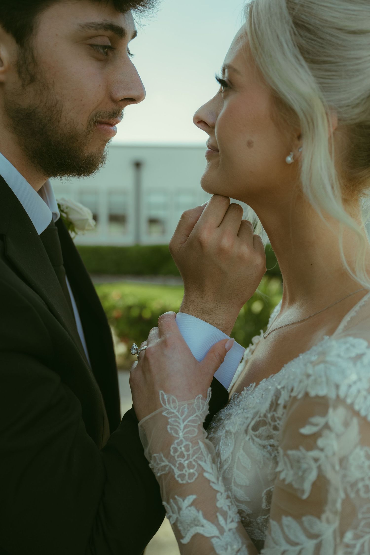 Groom tenderly lifts bride's chin as they gaze into each other's eyes during romantic wedding portrait outdoors.