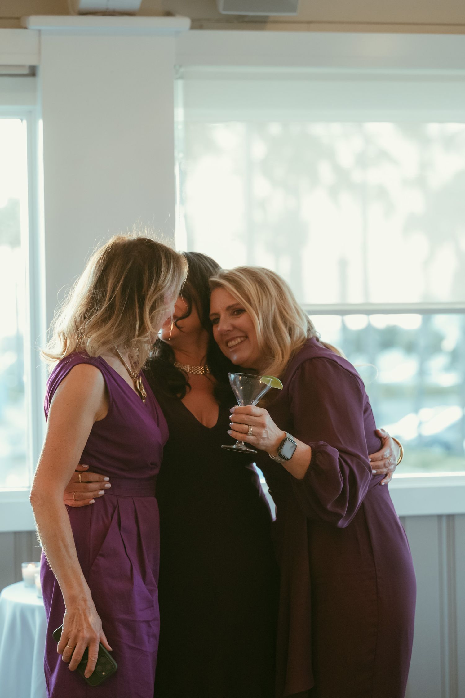 Three women in purple and black dresses share a warm embrace while holding cocktails at a wedding reception.