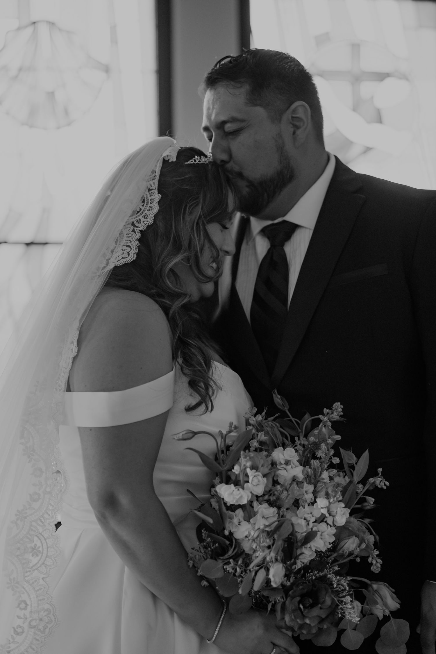 Black and white wedding photo of bride and groom sharing a kiss in church, bride holding floral bouquet.