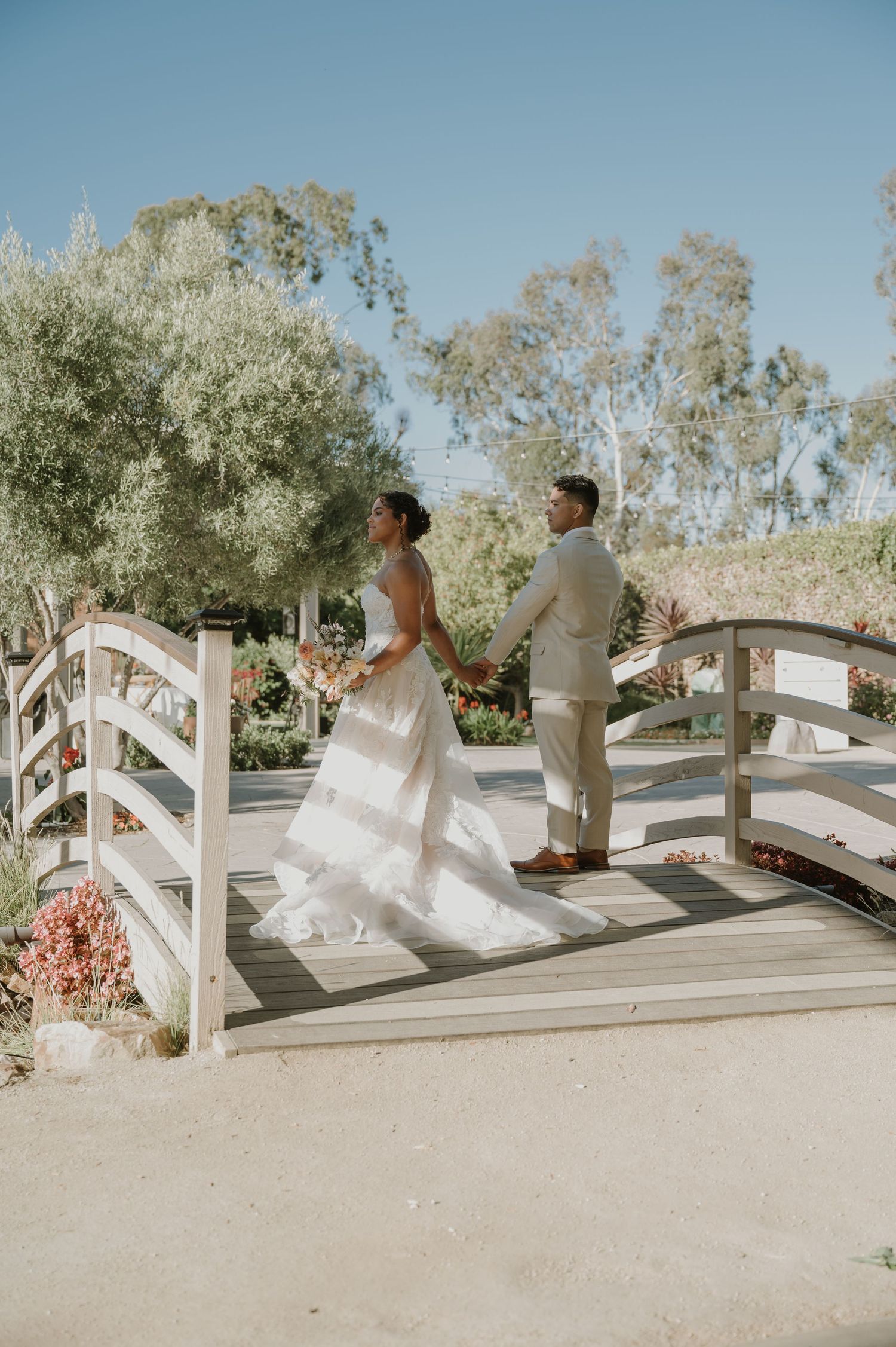 Bride and groom stand on a white wooden bridge surrounded by lush greenery on a sunny wedding day.