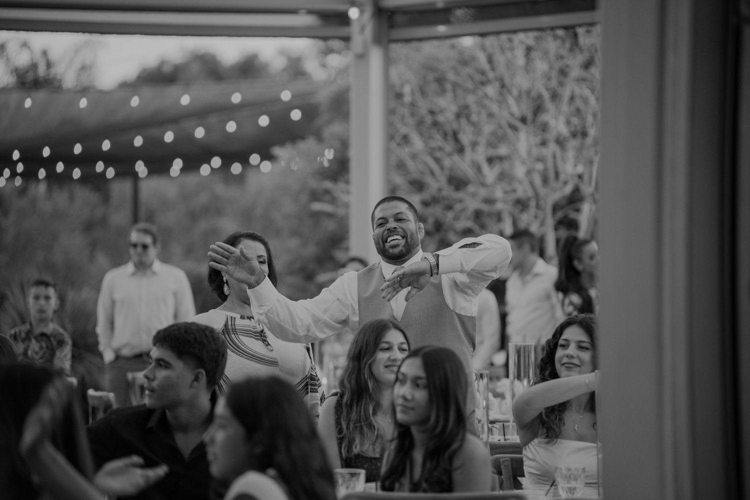 Wedding guests celebrate and cheer during reception under string lights in black and white photo.