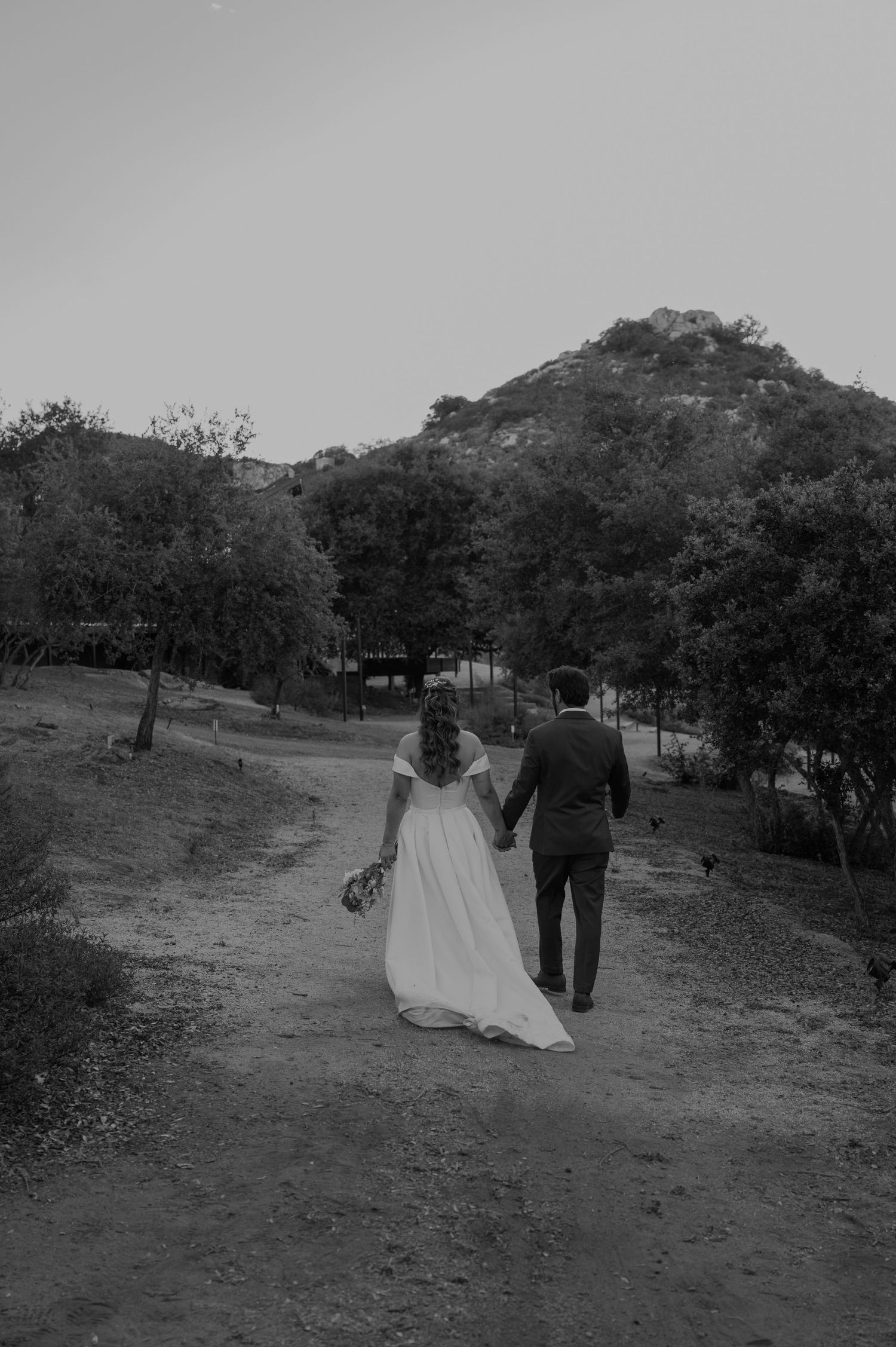 Bride and groom walking hand in hand down a dirt path with a mountain backdrop in black and white.