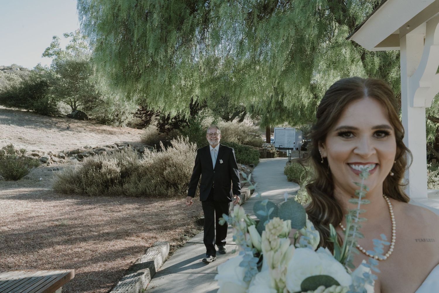 Bride smiling with bouquet as father walks toward her on garden path at outdoor wedding ceremony.