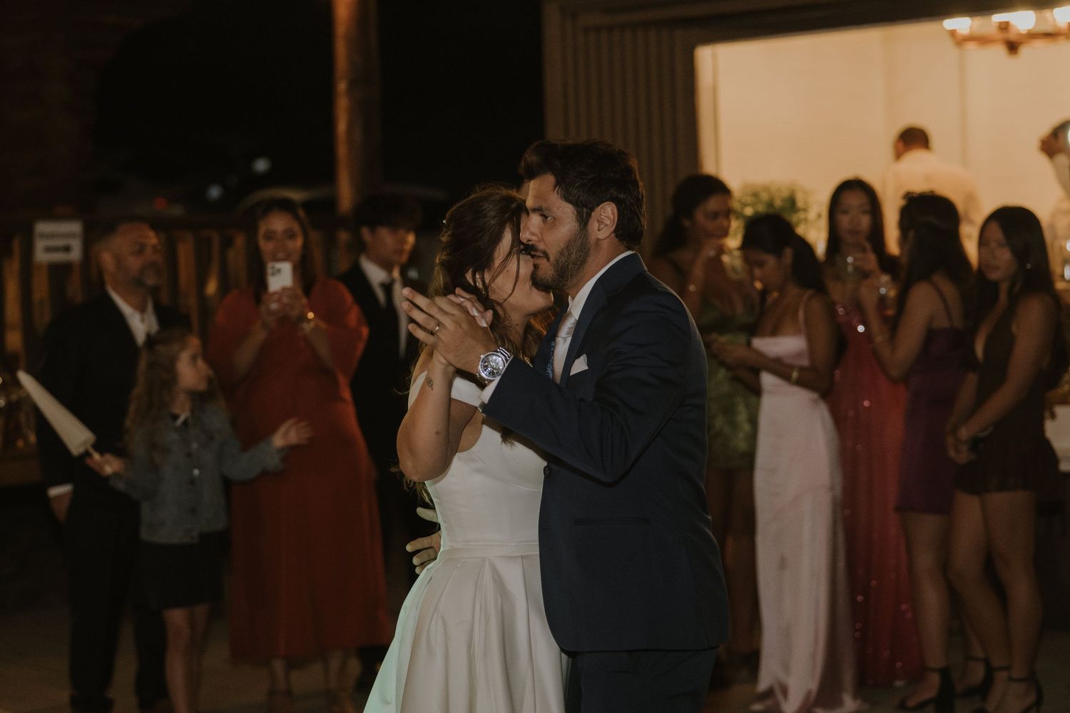 Bride and groom share first dance kiss at outdoor wedding reception with guests watching and photographing the moment.