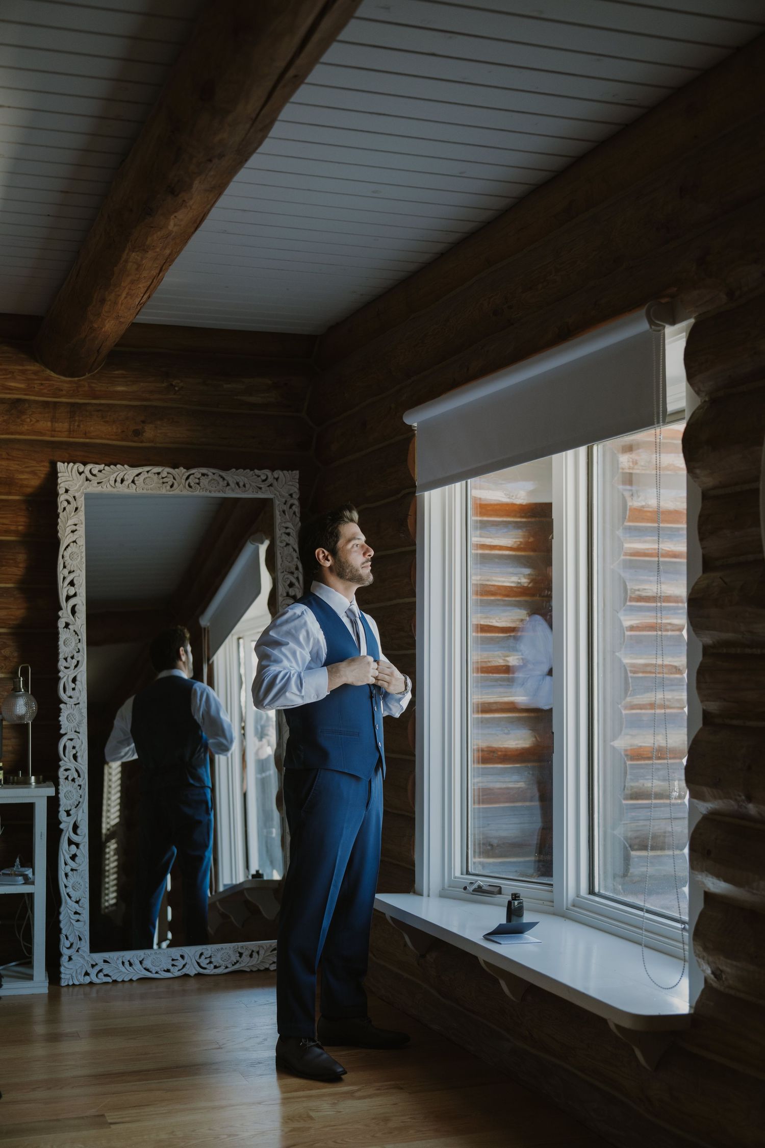 A groom in a blue suit adjusts his shirt near a window in a rustic log cabin room with a decorative mirror.