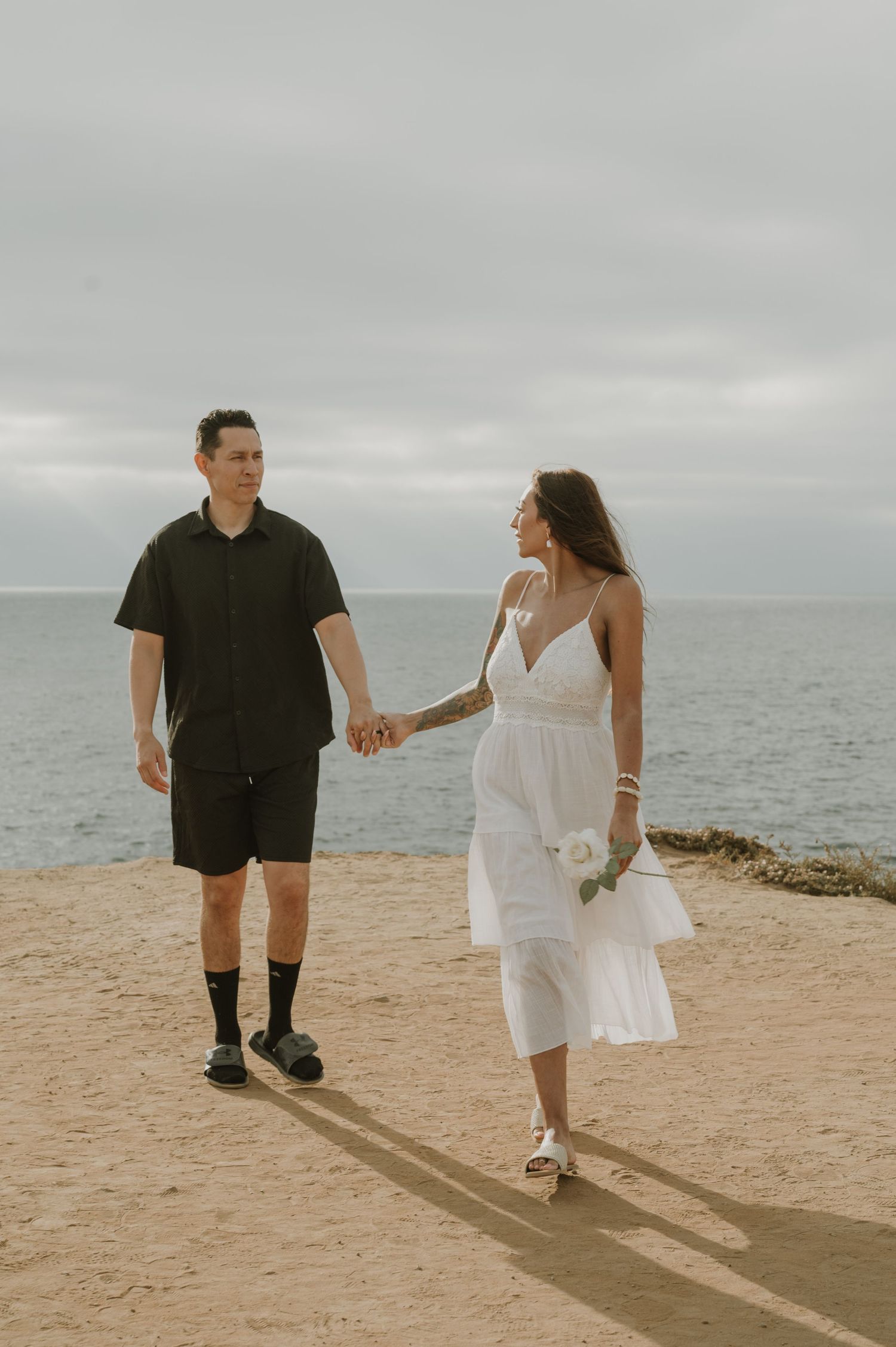 Couple in black and white outfits walking playfully along a sandy beach.