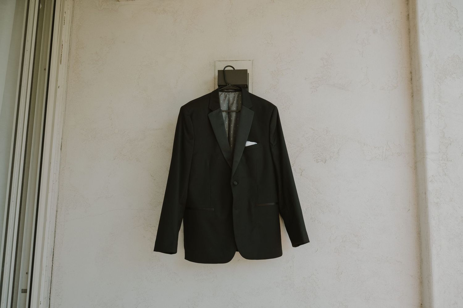 A black suit jacket hanging against a white wall in preparation for a wedding.