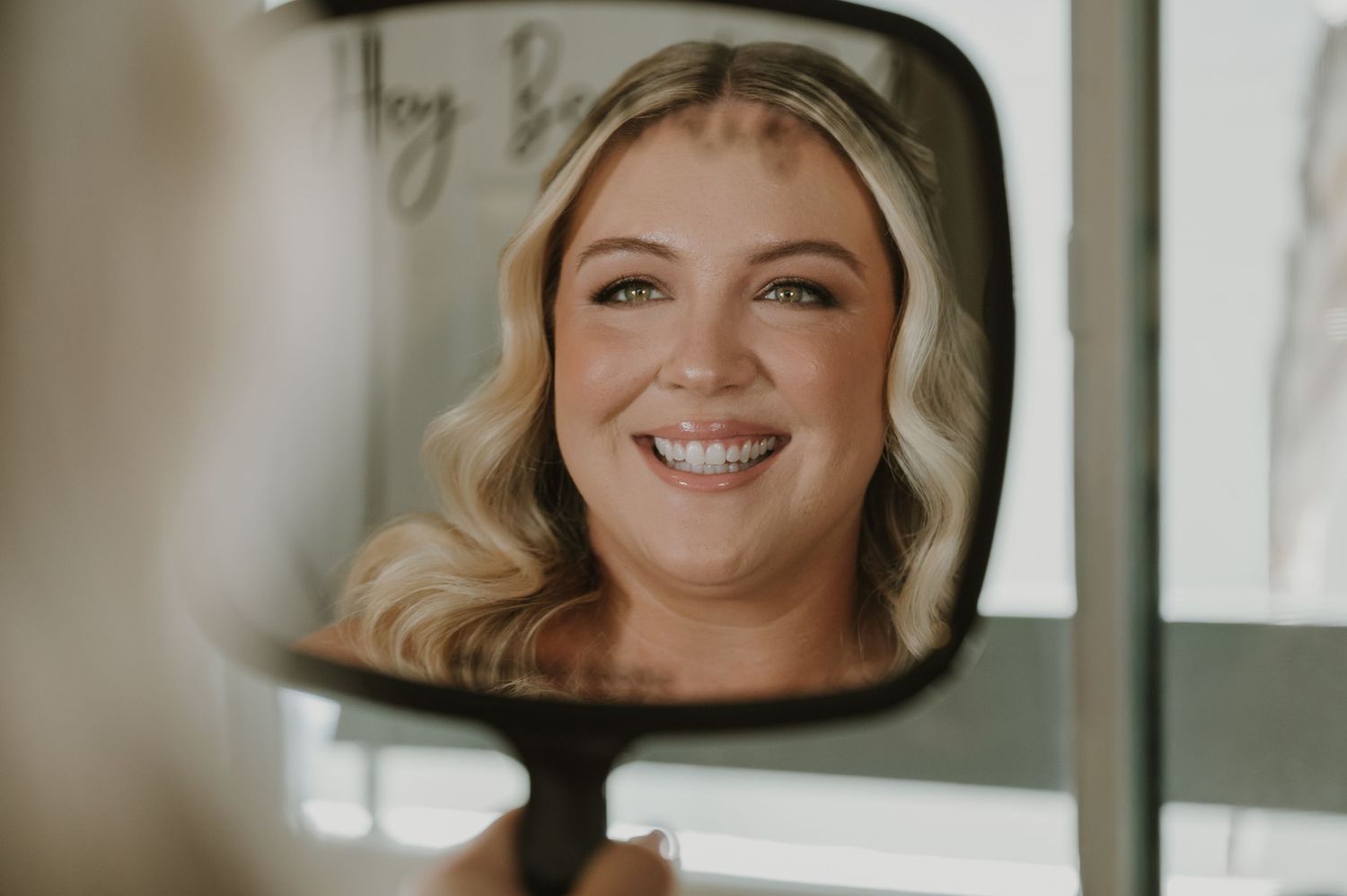 Bride smiling at her reflection in a handheld mirror, showcasing elegant makeup and soft blonde waves.