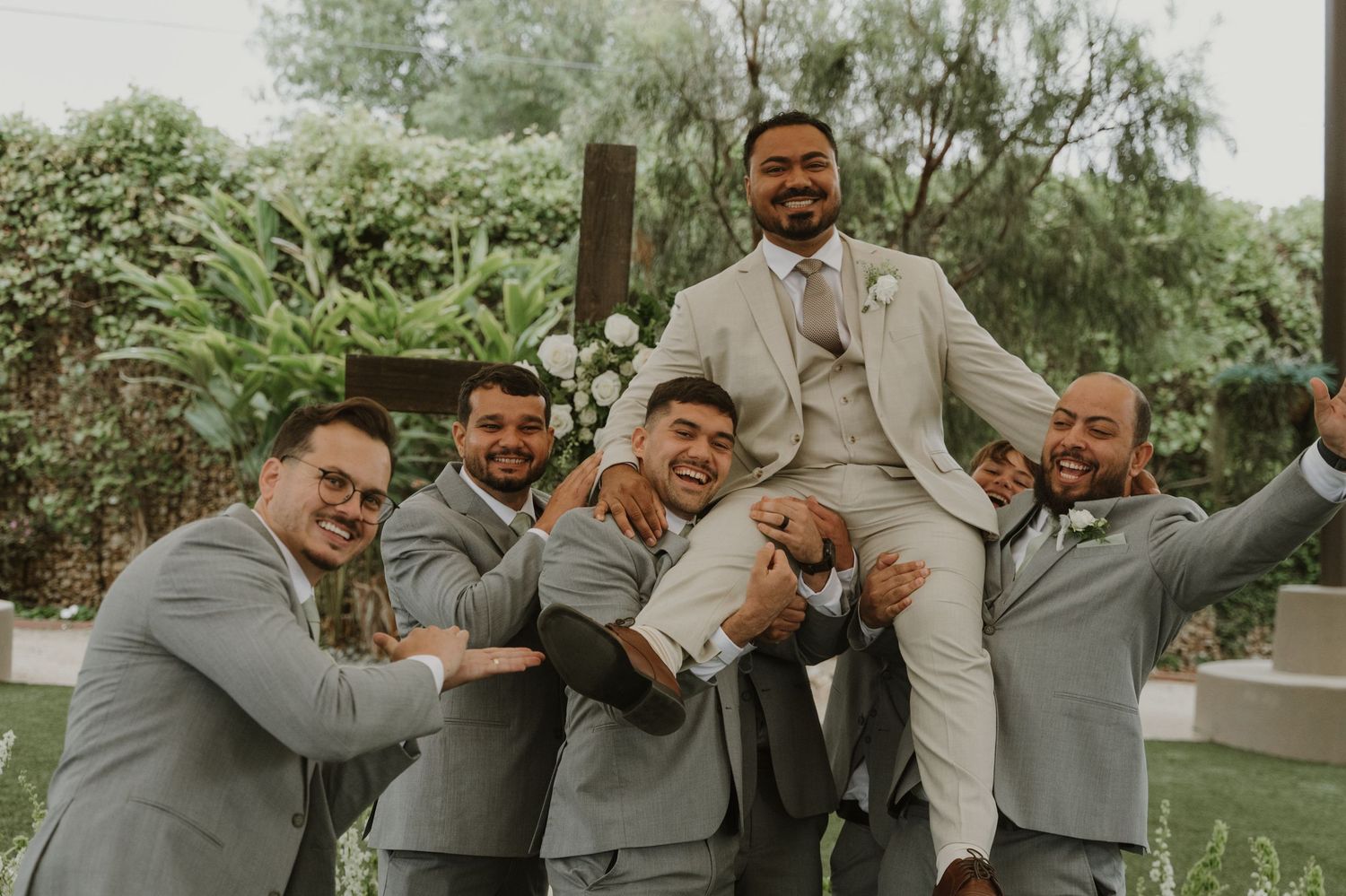 Groom in beige suit lifted by smiling groomsmen in gray suits at outdoor wedding ceremony.