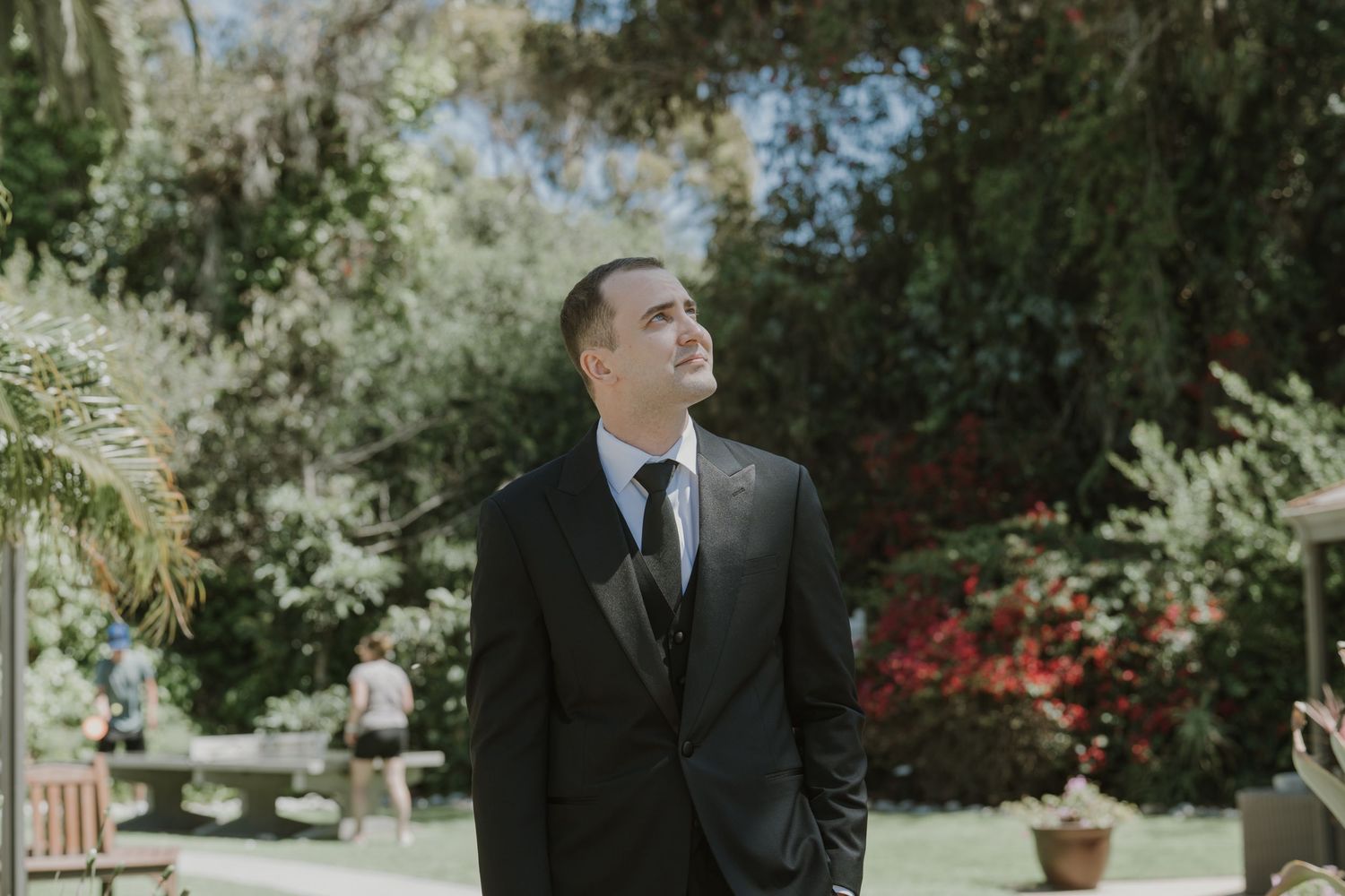 Groom in dark suit and tie looking up in lush garden setting on wedding day.