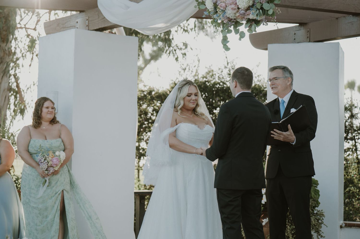 Bride and groom exchange vows at outdoor wedding ceremony beneath white floral arch with bridesmaid nearby.