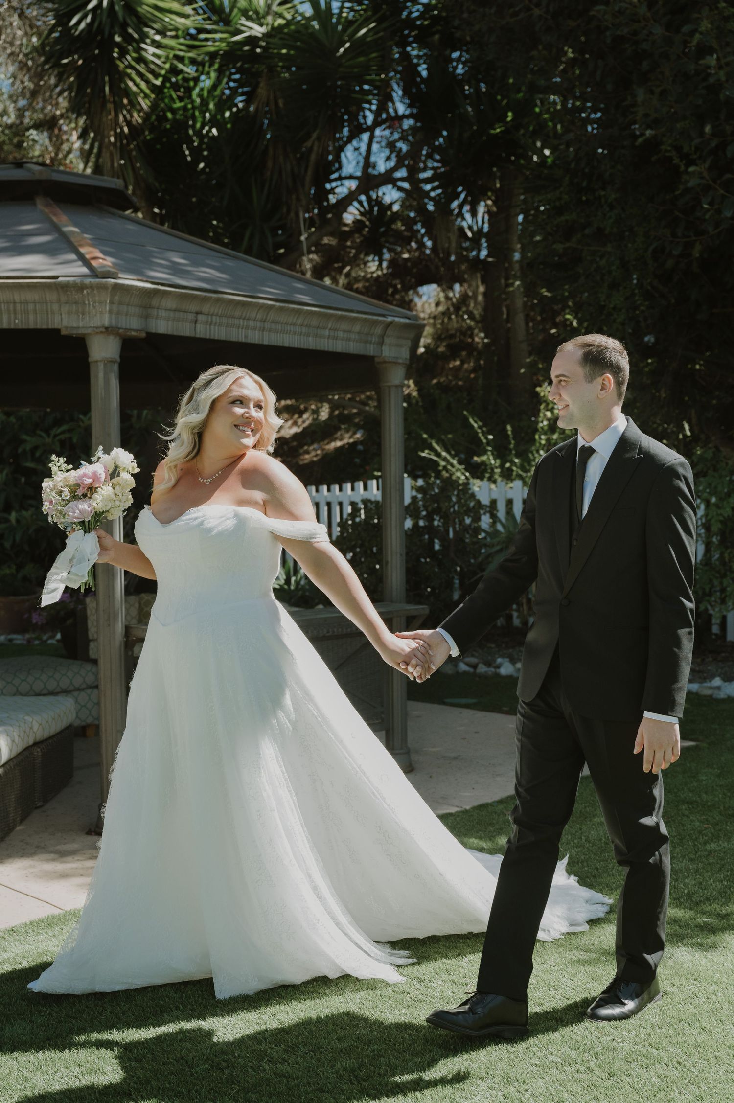 Bride in white gown holds bouquet while holding hands with groom in black suit in a garden gazebo setting.