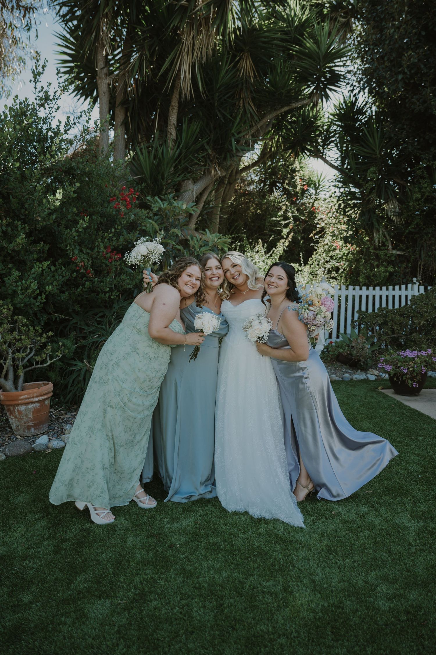 Bride in white gown poses with three bridesmaids in blue and green dresses holding bouquets in a garden.