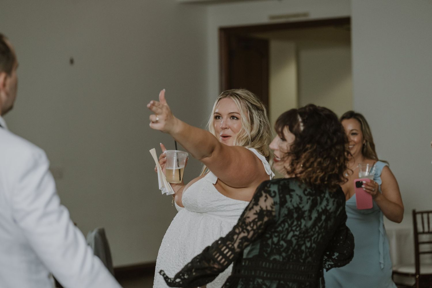 Bride in white dress laughing and dancing with guests while holding a drink at wedding reception.