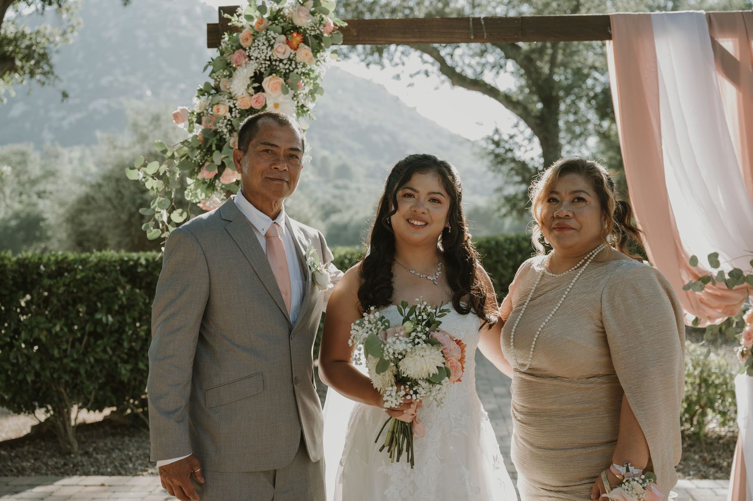 Bride poses with parents at outdoor wedding ceremony with floral arch and mountain backdrop.