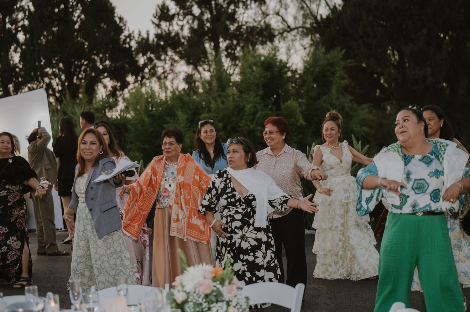 Women in colorful dresses celebrate at an outdoor wedding reception, laughing and dancing together at dusk.