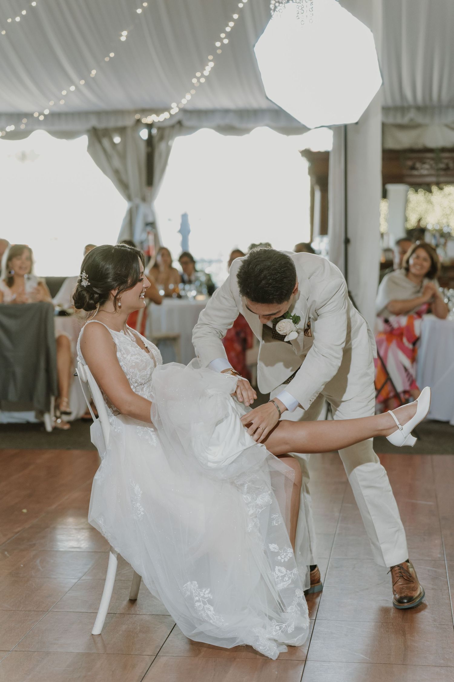 Groom in ivory suit removes bride's garter during wedding reception dance under string lights in a white tent.