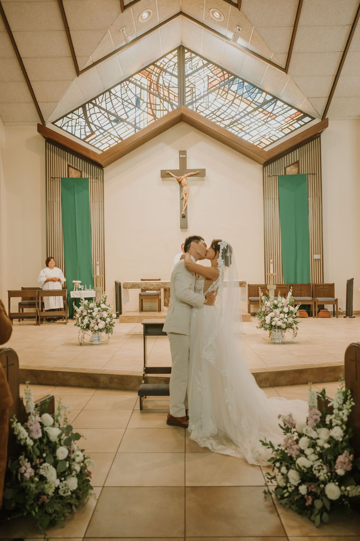 Bride and groom share first kiss at church altar adorned with floral arrangements and crucifix during wedding ceremony.