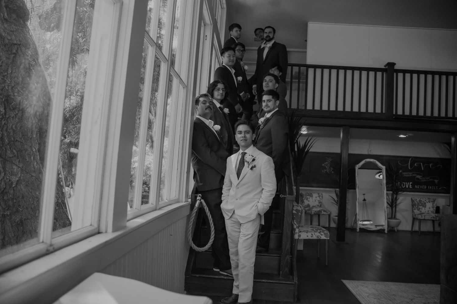 Groom in white suit poses with groomsmen on staircase in black and white wedding photo.