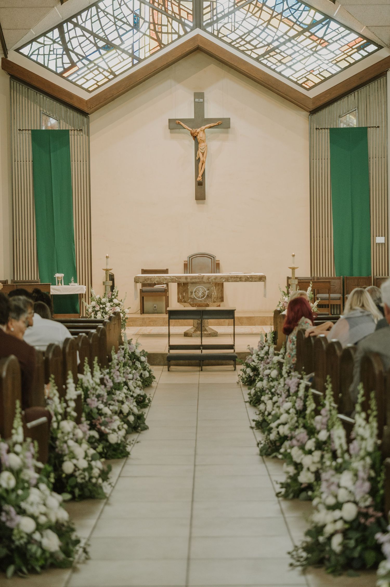 Catholic church wedding aisle adorned with white floral arrangements leading to altar with crucifix and green banners.