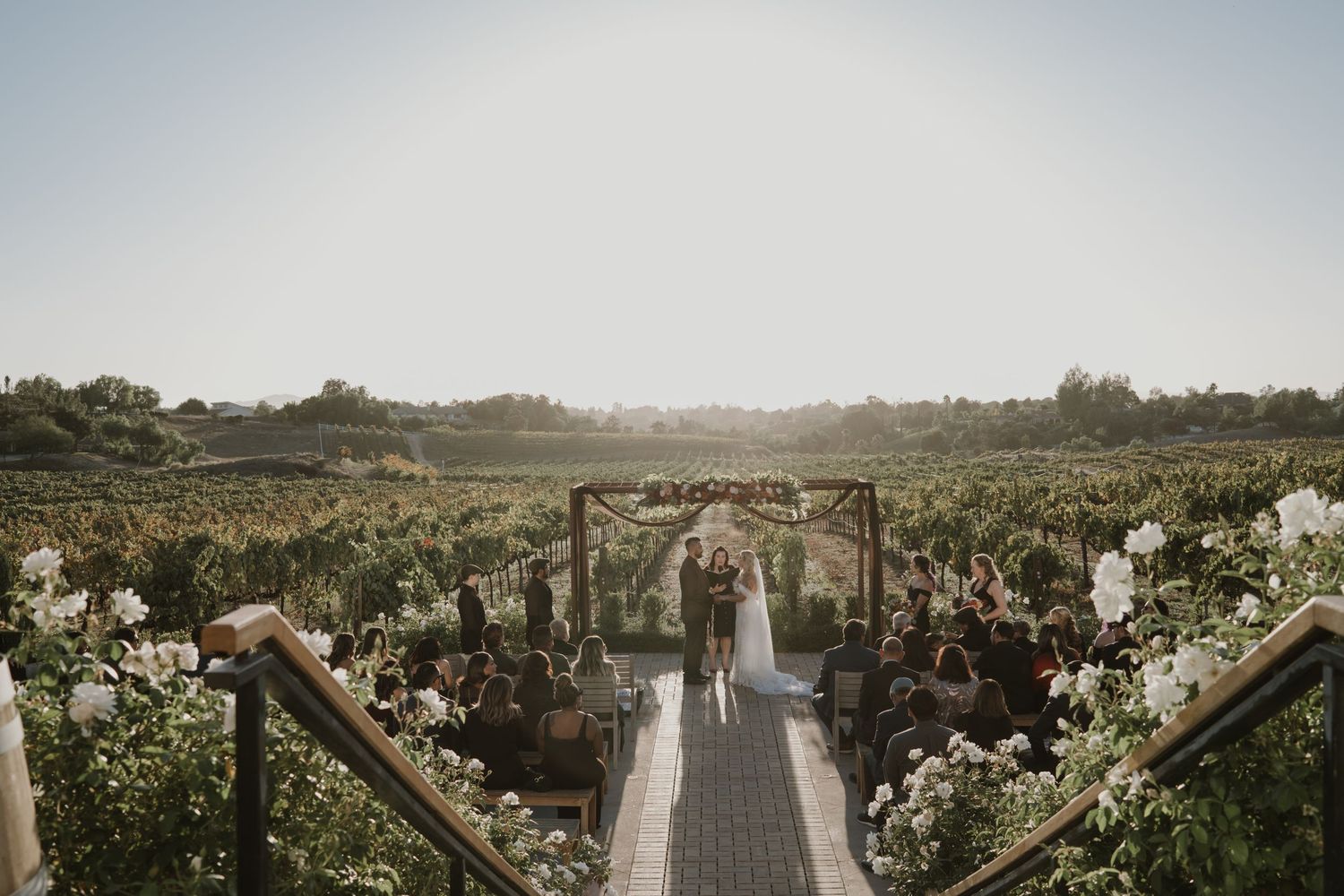 Aerial view of a vineyard wedding ceremony setup with white chairs and floral decorations along a pathway.