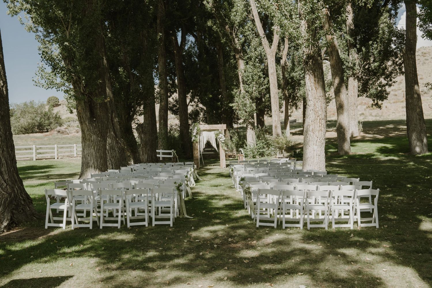 White folding chairs arranged in rows for an outdoor wedding ceremony beneath tall trees.