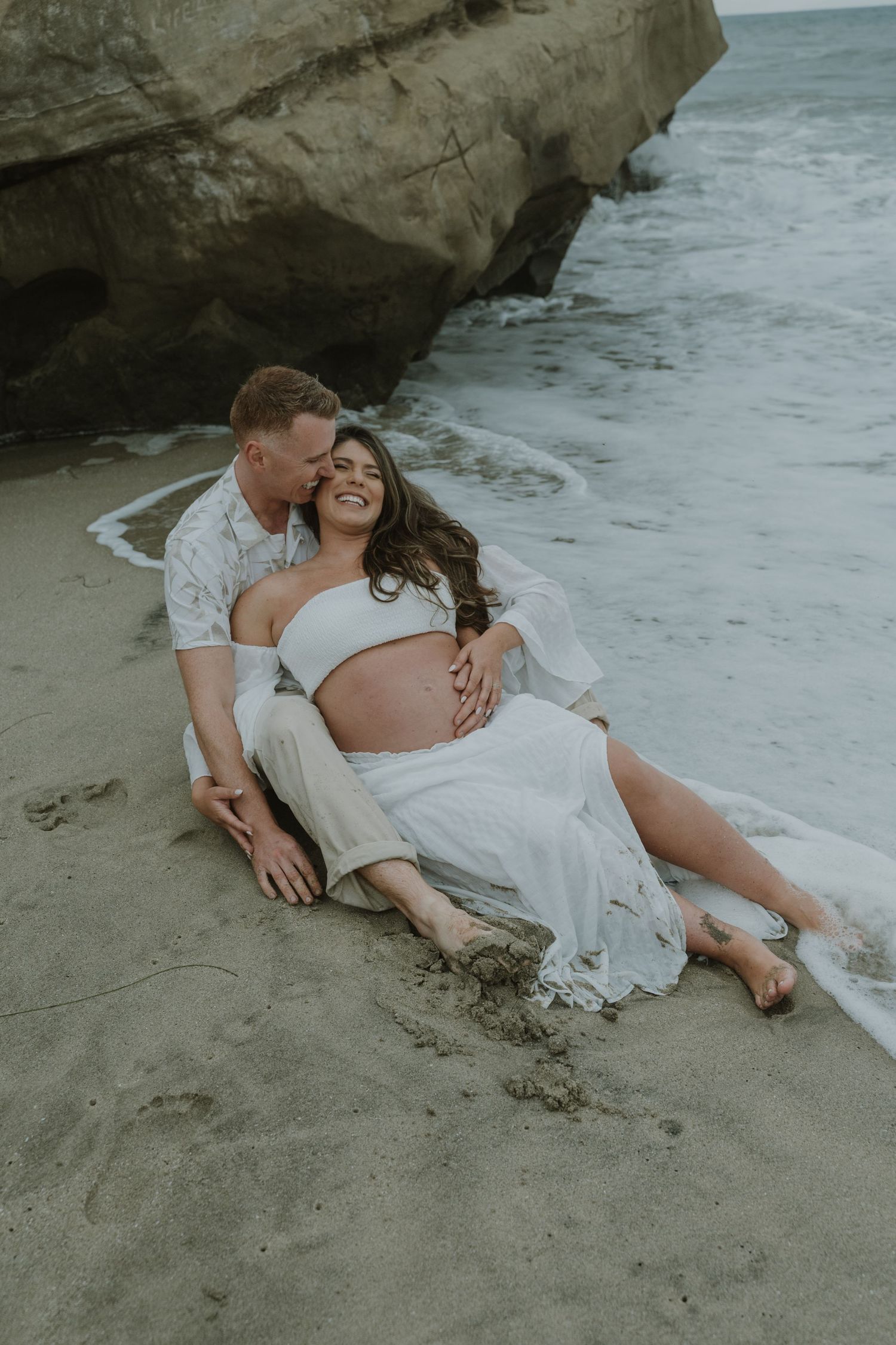 Tender maternity beach photos with couple resting on sandy shore near rock formations.