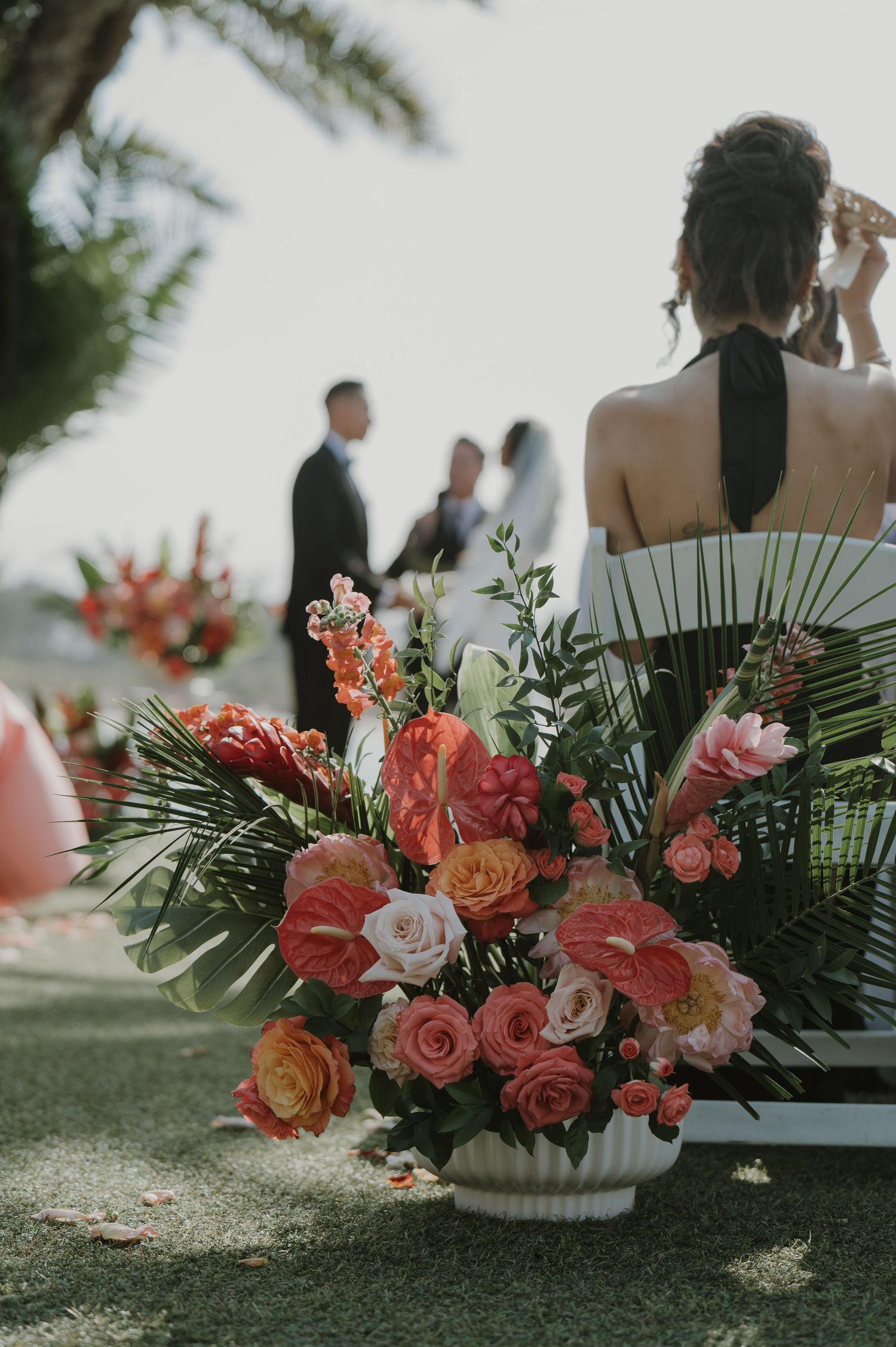 Coral and pink floral arrangement with palm leaves at outdoor wedding ceremony.