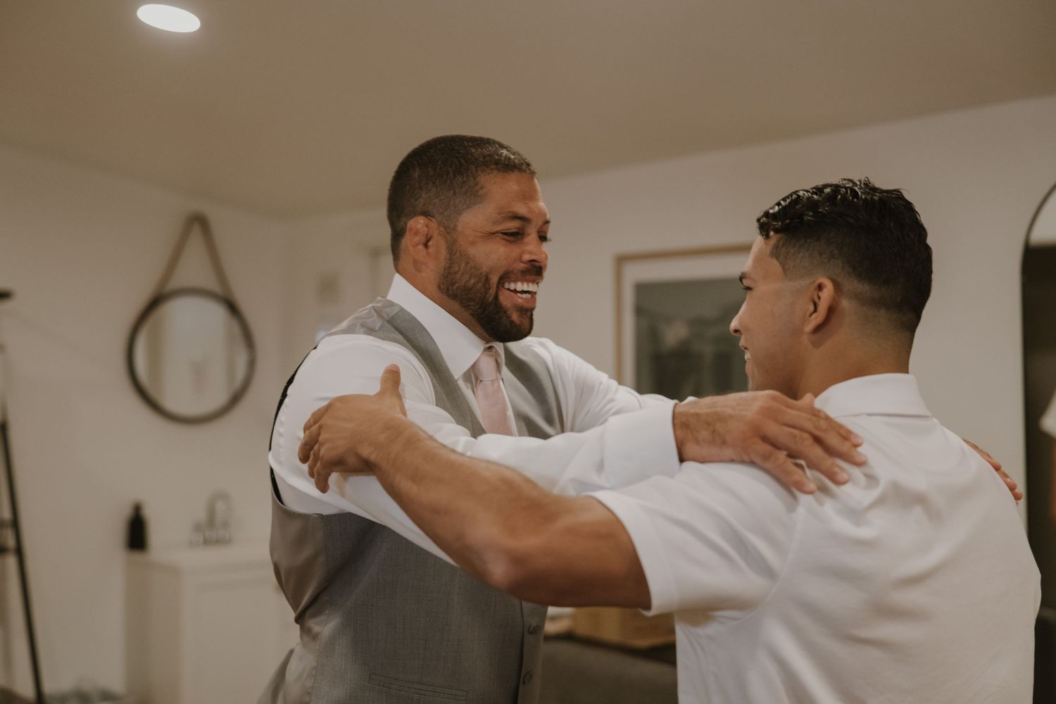 Two groomsmen share a joyful embrace and smile during wedding day preparations indoors.
