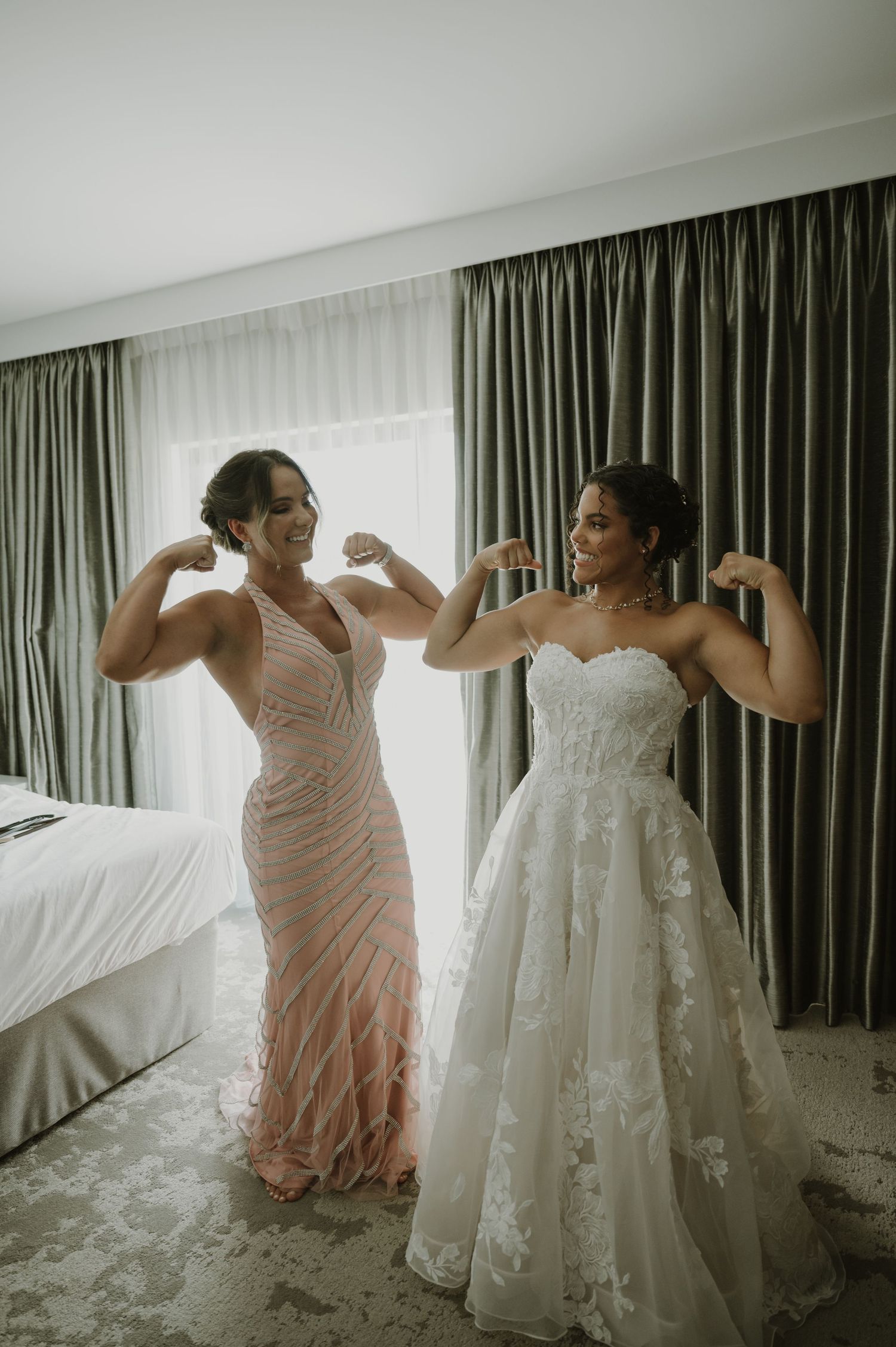 Bride in lace wedding gown and bridesmaid in pink dress flexing muscles together in hotel room.