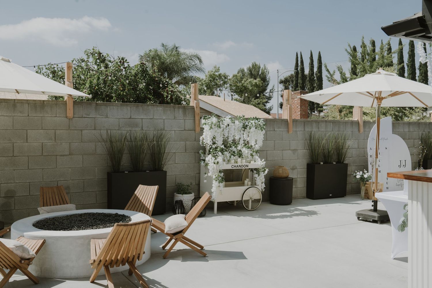 Modern outdoor patio with fire pit, teak chairs, white umbrellas, and lush greenery against a concrete block wall.