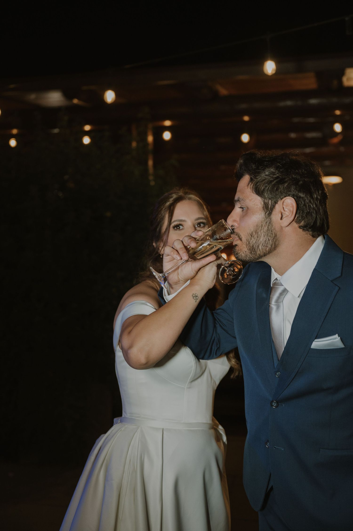 Bride and groom sharing a romantic champagne toast kiss at outdoor wedding reception at night.