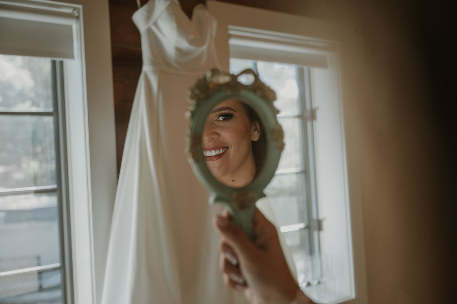 Smiling bride admiring her reflection in a vintage hand mirror before her wedding.