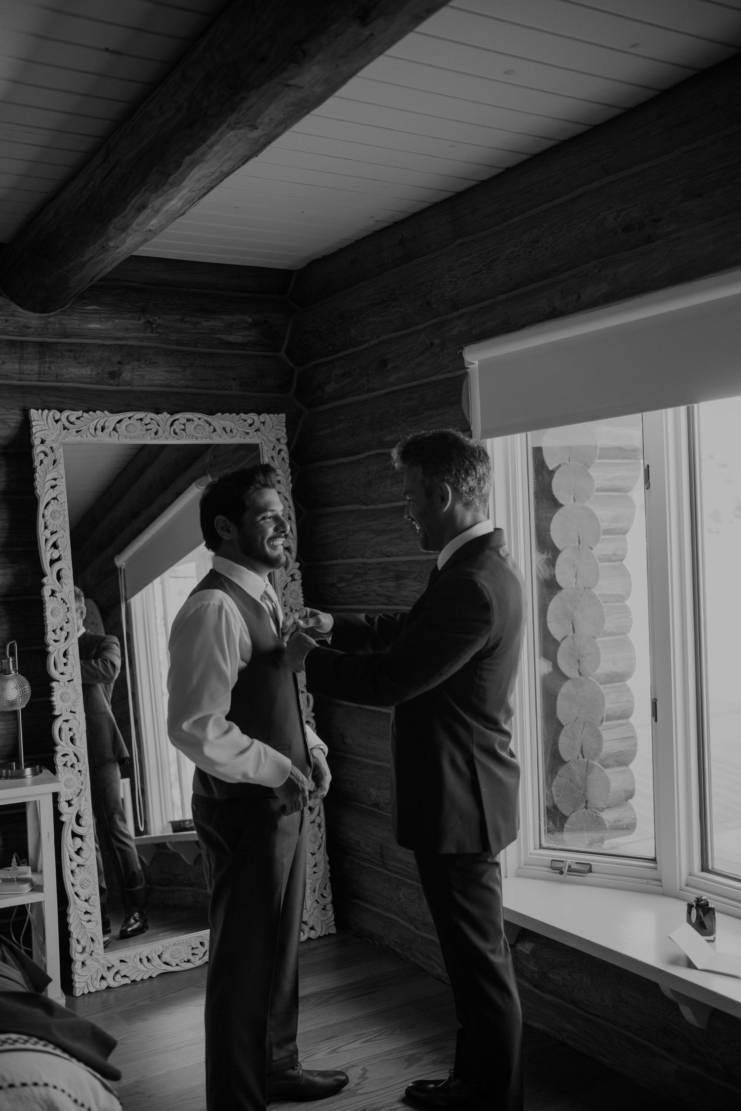 Groom getting his tie adjusted by a groomsman in a rustic log cabin room with a decorative mirror.