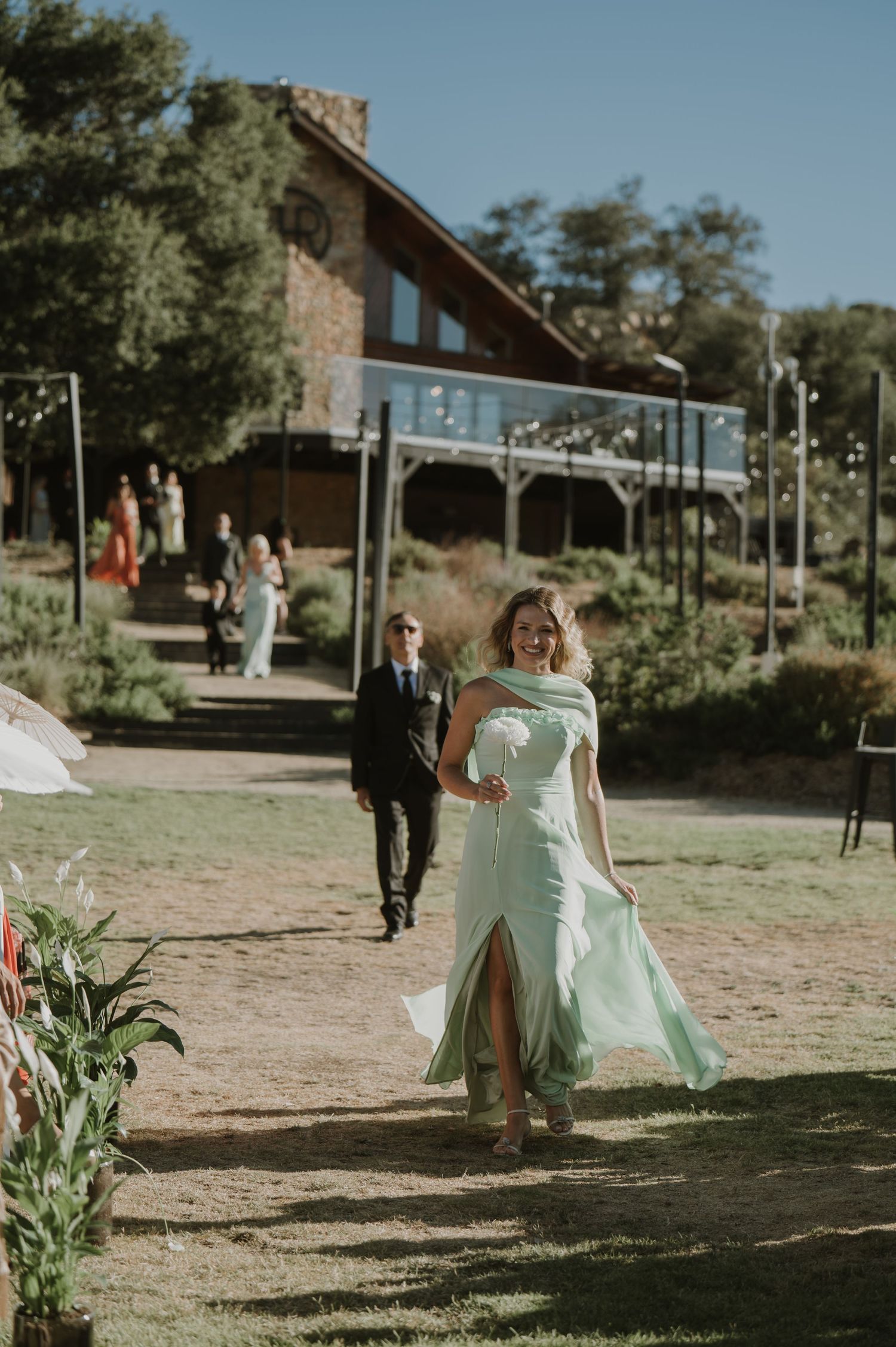 Woman in mint green dress walks toward camera at outdoor wedding venue with rustic lodge building in background.