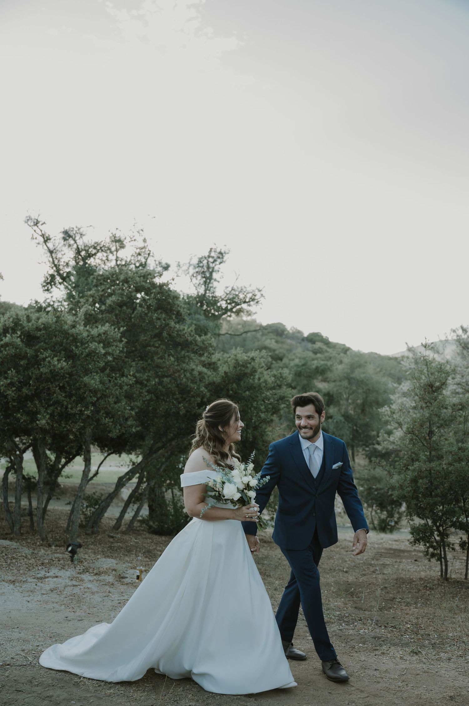 Bride in white off-shoulder gown and groom in navy suit walk together outdoors among green trees on their wedding day.