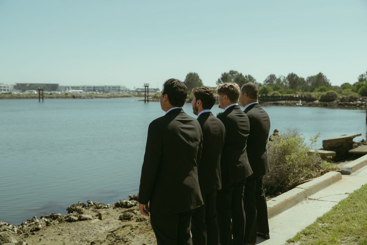 Four groomsmen in dark suits standing together by a waterfront, gazing out over the calm blue water on a sunny day.