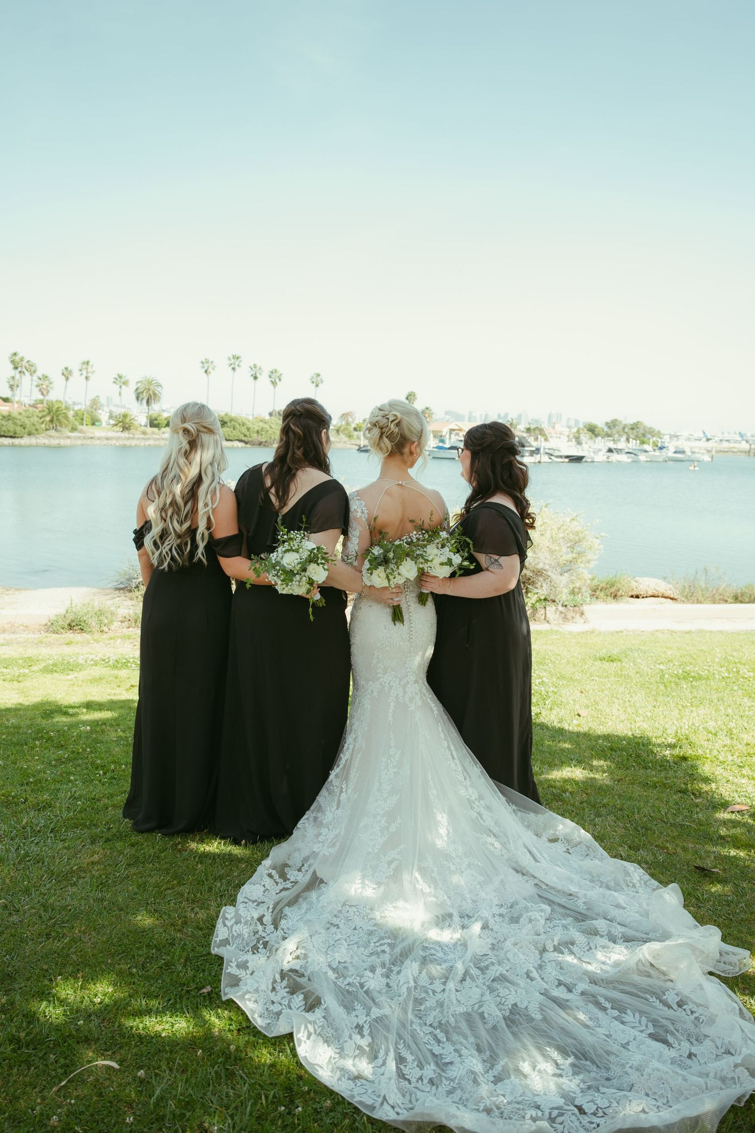 Bride in lace gown with bridesmaids in black dresses holding bouquets by a waterfront.