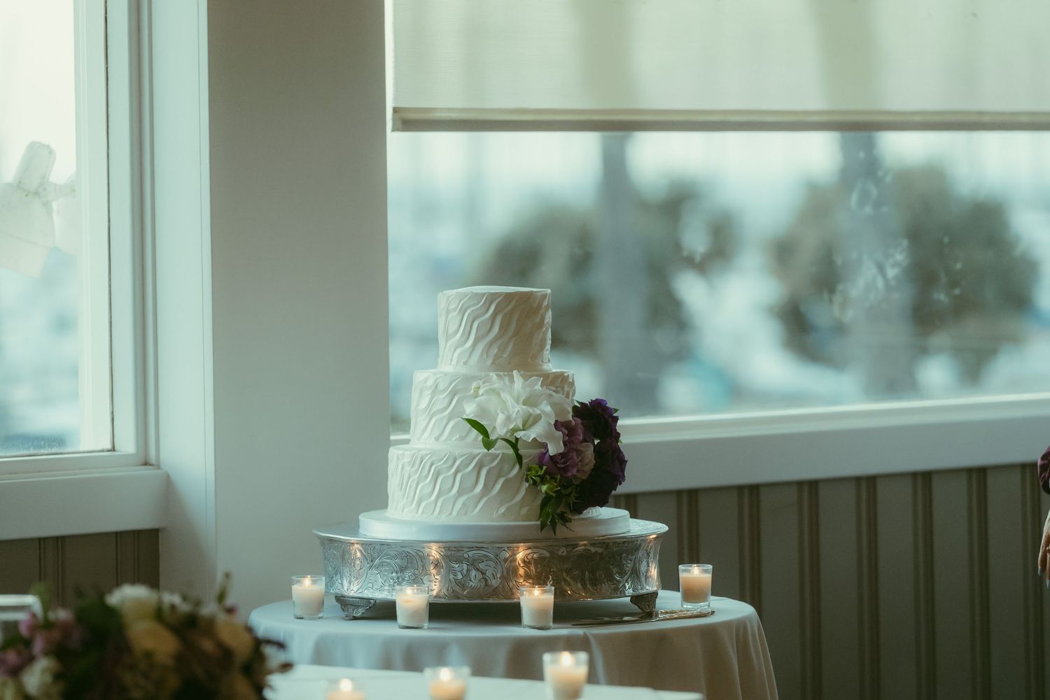 Elegant three-tier white wedding cake adorned with purple flowers displayed near window with candles.