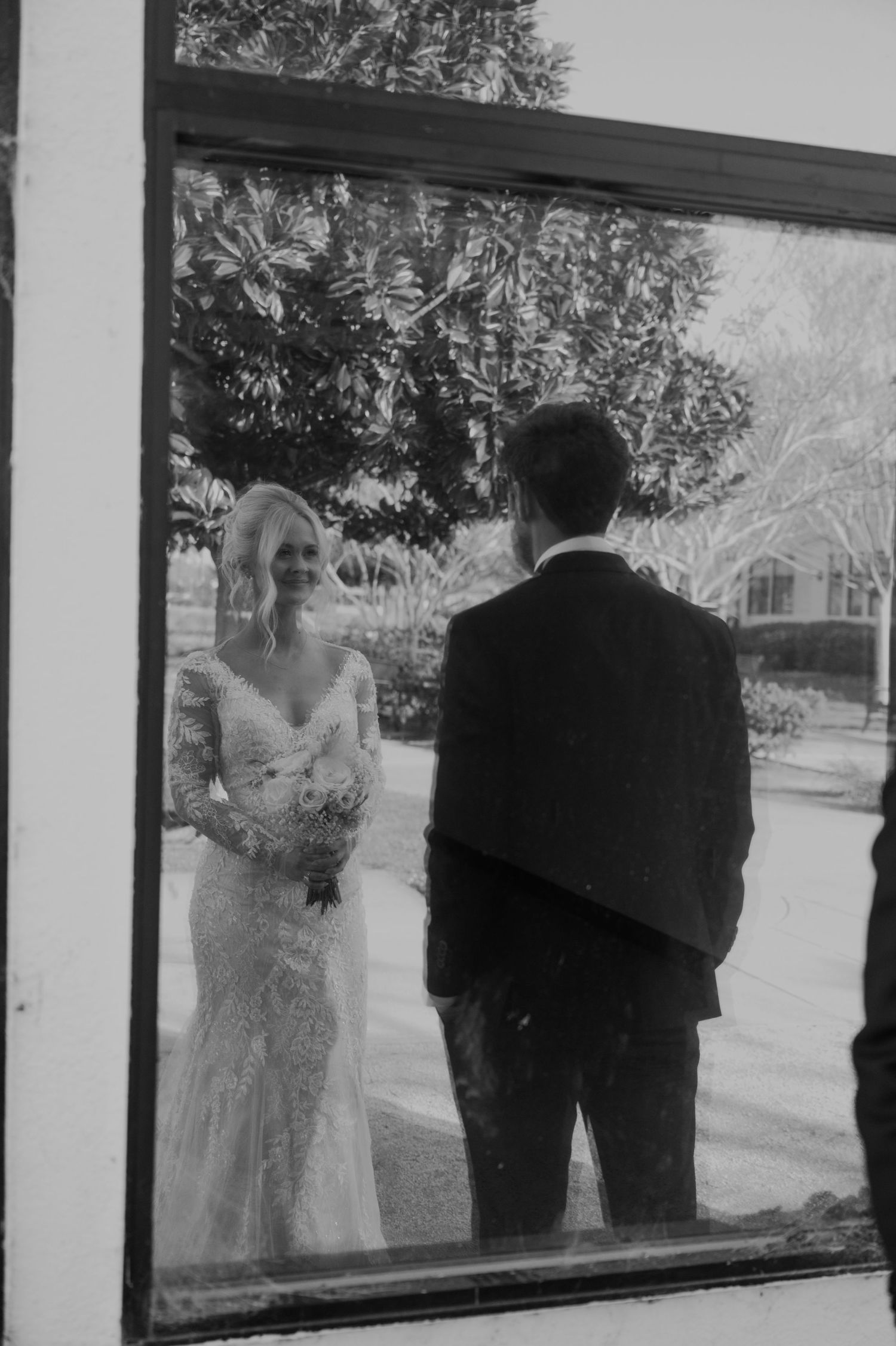 Black and white wedding photo of bride in lace gown and groom in suit reflected in a window outdoors.