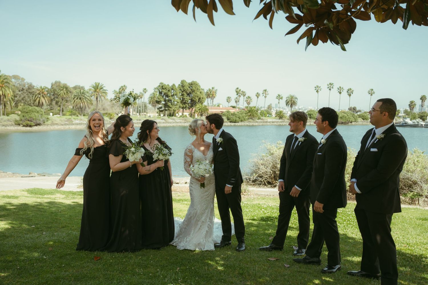 Wedding party in black attire poses by a scenic lake, with bride and groom sharing a kiss surrounded by bridesmaids and groomsmen.