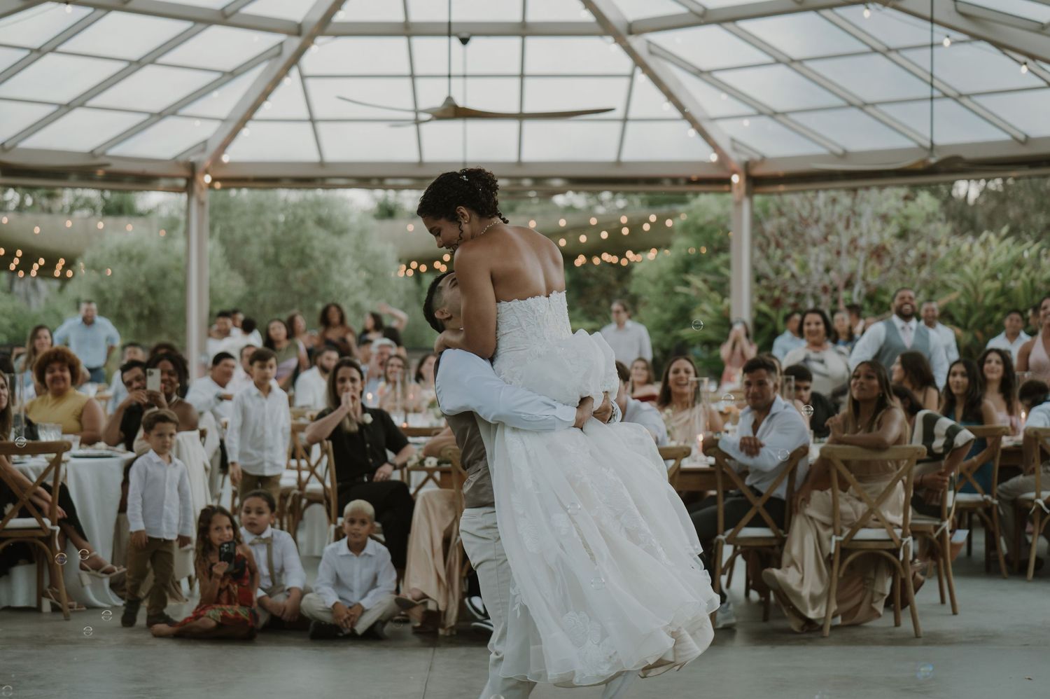Couple shares first dance in elegant outdoor wedding reception under glass pavilion with string lights.
