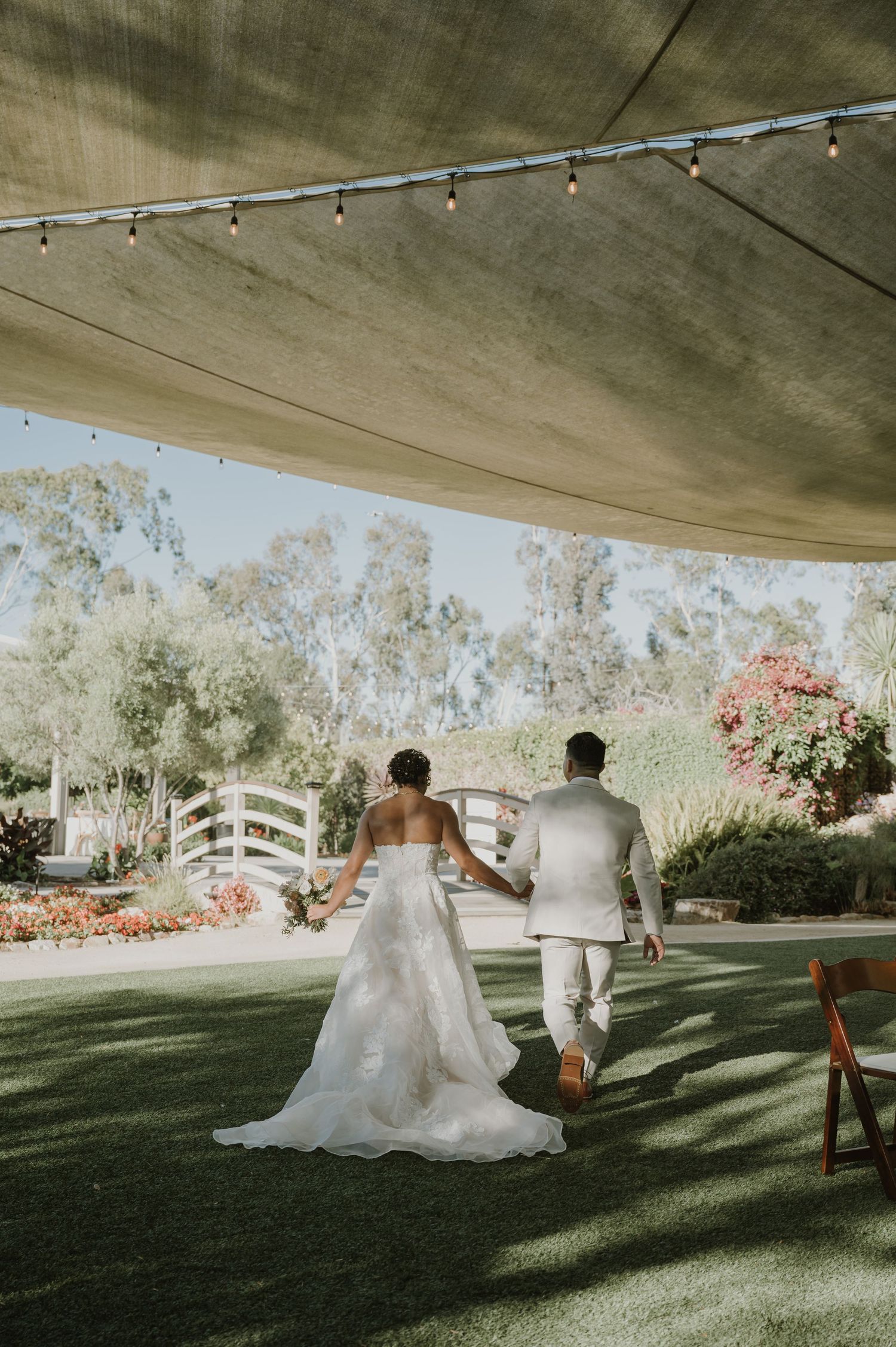 Bride and groom walking hand in hand across a lush garden wedding venue under a draped tent.