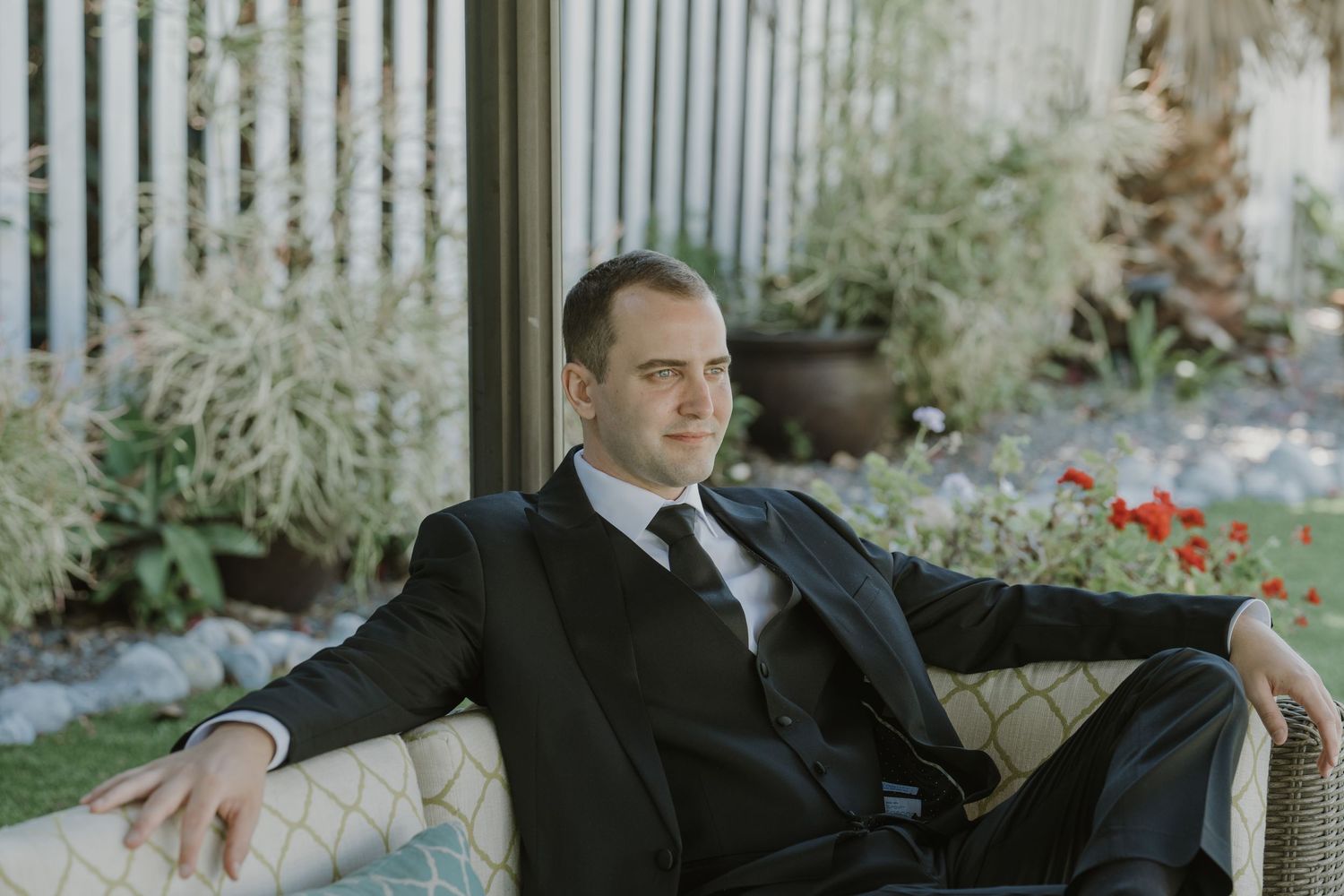Man in black suit sitting relaxed on outdoor sofa with garden background.